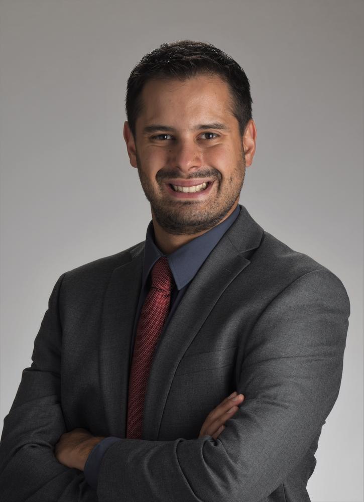 smiling man with arms crossed wearing gray jacket, dark blue shirt and red tie