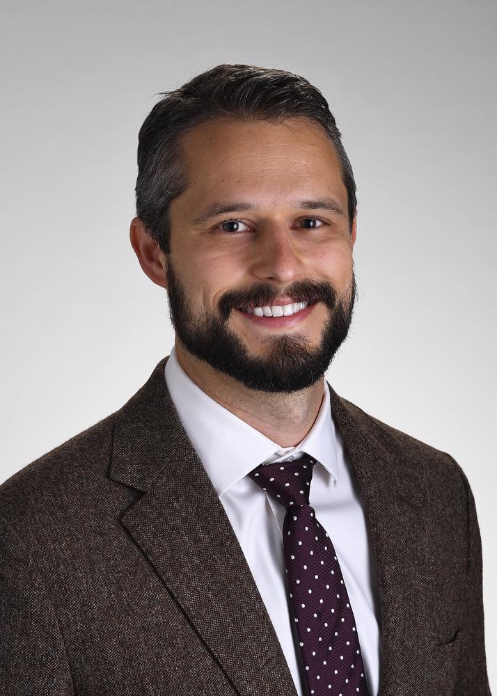 Smiling man with beard. He is wearing a brown suit jacket, white button down shirt and a brown tie with white dots.