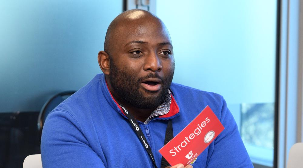 A man wearing a blue pullover with a lanyard around his neck. He is holding a red card that says strategies.