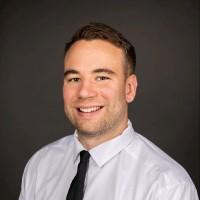 photo of Aaron Smith smiling; he is wearing a white shirt with a black tie