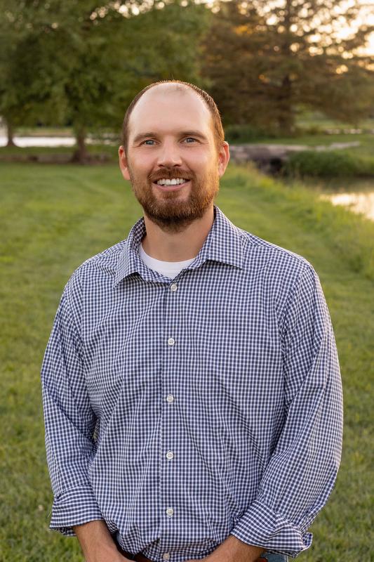 smiling man standing near a lake in a white and blue checked shirt