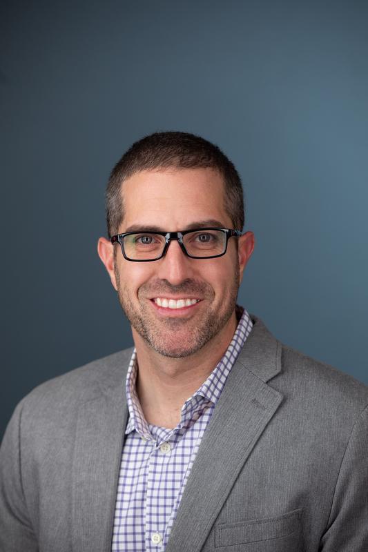 smiling man in gray jacket, white a blue checked shirt and glasses