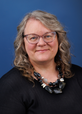 smiling woman in black wearing glasses and large black necklaces in front of blue background