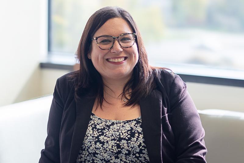 smiling woman in black jacket and black and white shirt wearing glasses in front of a window