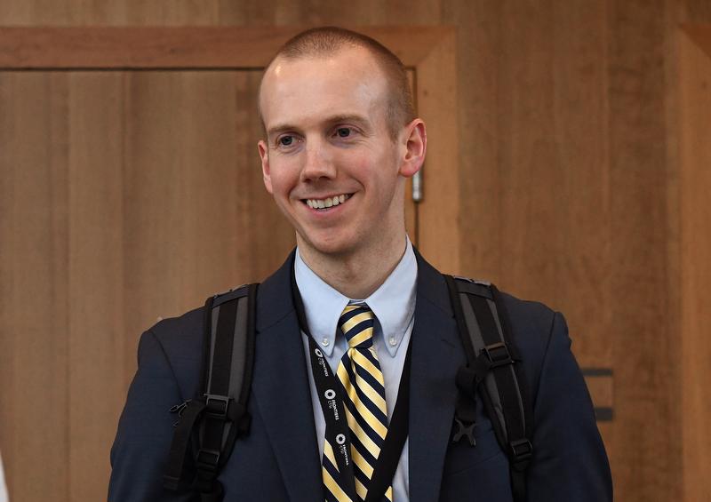 man smiling wearing black jacket, blue shirt and yellow and black tie with a backpack
