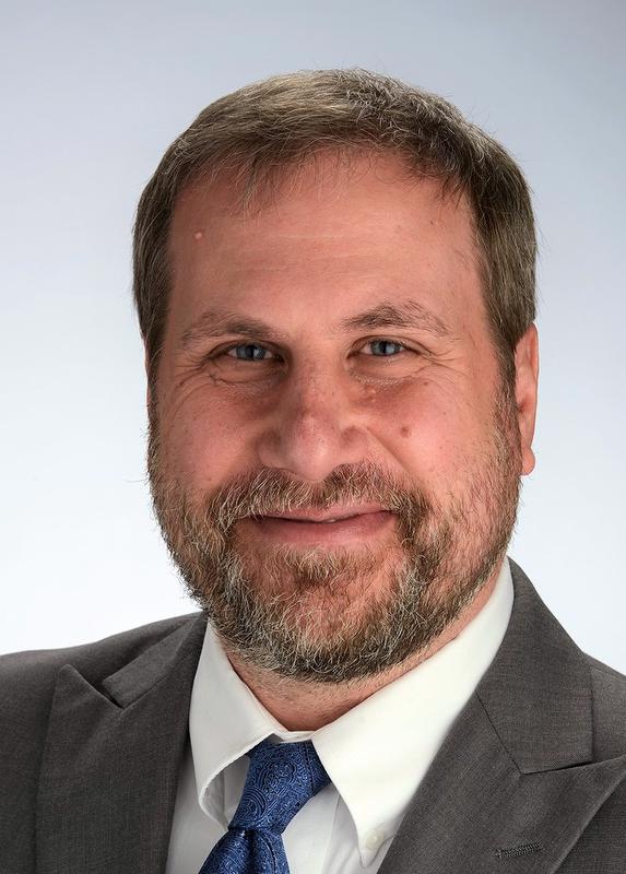 smiling man with beard wearing a brownish gray jacket, white shirt and blue tie