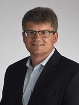 smiling man wearing glasses in black jacket and blueish button down shirt