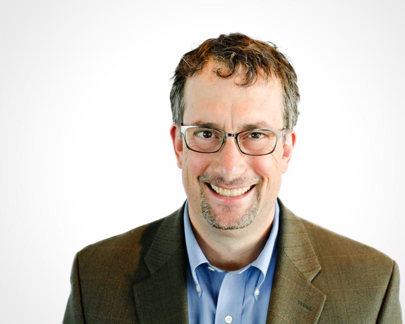 smiling man wearing glasses, a brown jacket and blue shirt with two buttons visible. also has a goatee