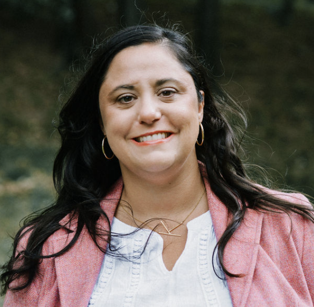 smiling person wearing pink jacket, white shirt, gold necklace and earrings
