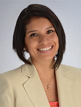 Smiling woman with longer hair in front wearing a beige jacket, pink shirt and necklace