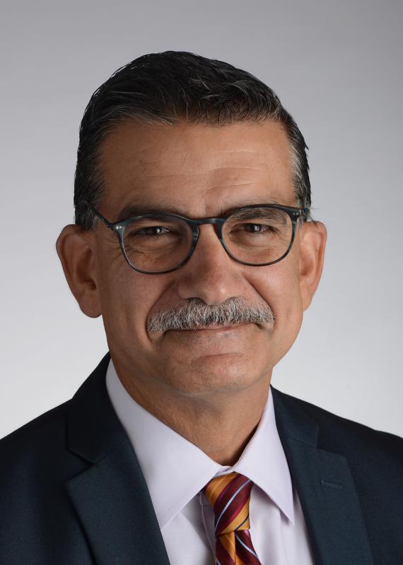 smiling man wearing a black jacket, white shirt and light orange tie with red, white and black stripes. also earing glasses
