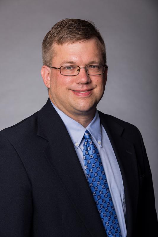 smiling man wearing a black jacket, blue shirt and blue tie with a pattern. also wearing glasses