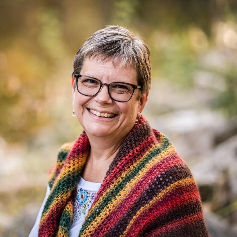 smiling woman with short hair wearing glasses, earrings a white shirt with designs at the neck and a multicolored sweater over her shoulders