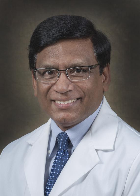 smiling man wearing a white doctor coat, blue shirt and blue and white checked shirt and glasses