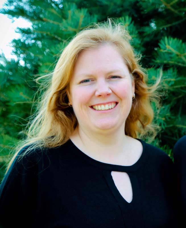 smiling woman with red hair wearing a black shirt with a fir tree behind her