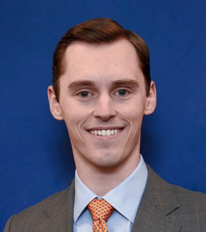 smiling man in front of a blue background wearing a dark gray jacket, blue shirt and orange tie with blue designs