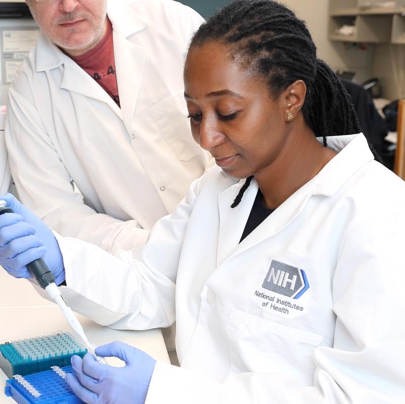 Woman in white lab coat wearing blue gloves holding a pipette in her right hand and touching the bottom of the pipette with her left. Tubes are in a case on the table in front of her and a person in a white lab coat watches