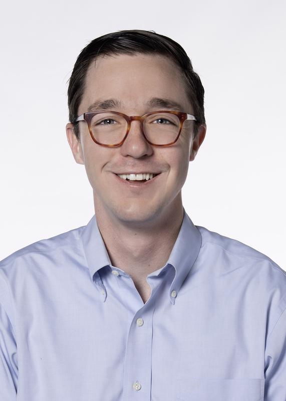 Smiling man in blue button up shirt and wearing reddish brown glasses.