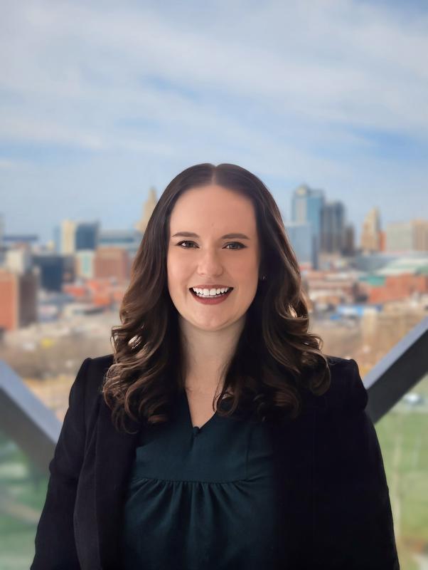 A smiling woman in a black suit jacket and dark colored shirt standing in front of a window that has a city landscape behind her.