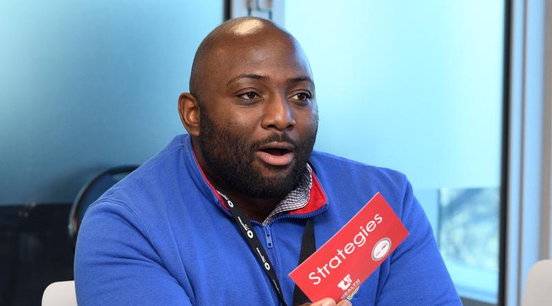 A man wearing a blue pullover with a lanyard around his neck. He is holding a red card that says strategies.