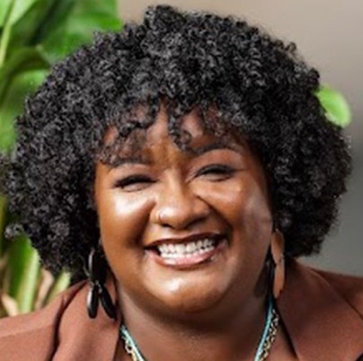 Smiling woman with short dark curly hair with a plant in the background. She is wearing a brown top, necklaces and two large earrings.