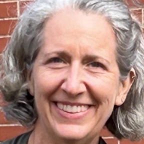 Smiling woman with chin-length gray hair. She is standing in front of a red brick wall.