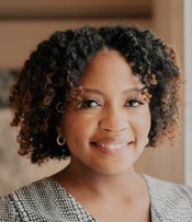 Smiling woman with chin length, dark curly hair. She is wearing earings and a white and black checked shirt.