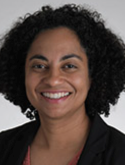 Smiling woman with dark curly hair. She is wearing a black suit jacket and darker pink shirt.