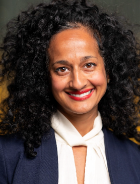 Smiling woman with below shoulder-length dark curly hair. She is wearing a blue suit jacket and a white shirt.