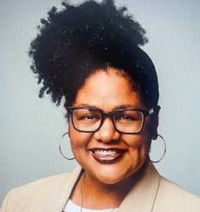 Smiling woman with dark hair that is in an updo. She is wearing a tan jacket, white shirt, big hoop earrings and glasses.